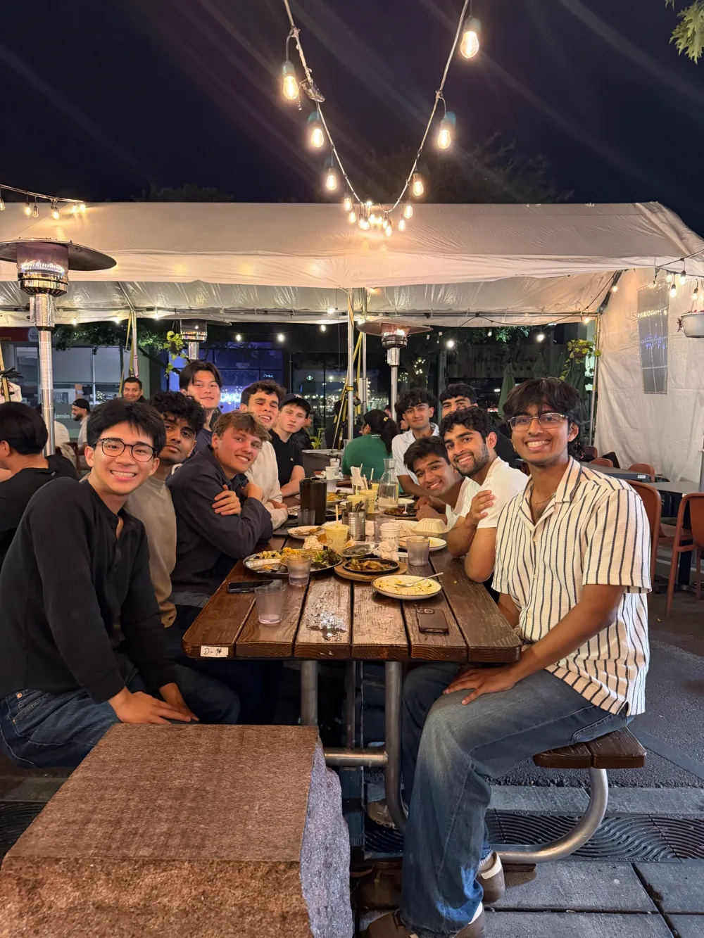 A long outdoor dinner table under string lights, lined with people smiling at the camera